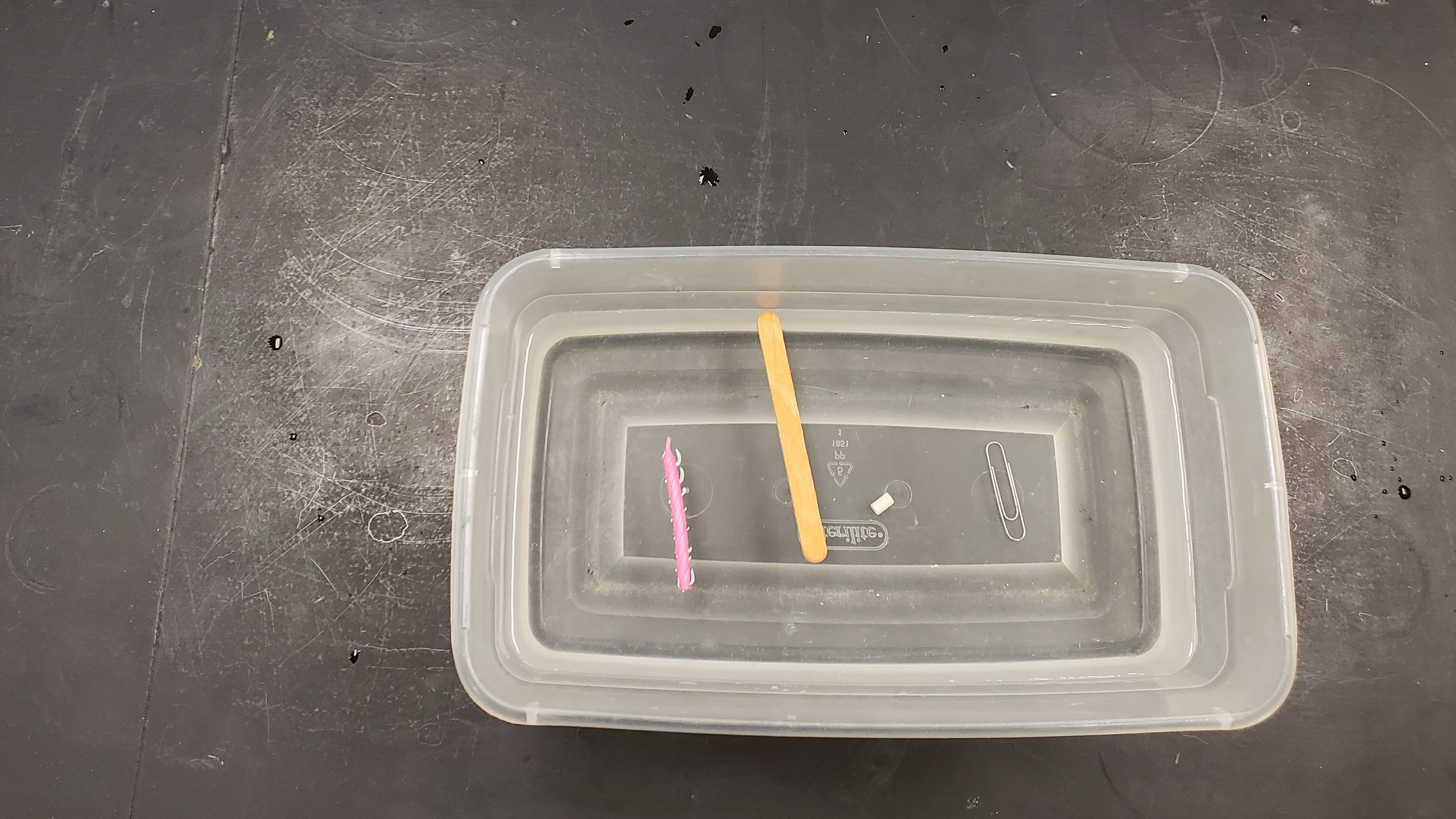 container filled with water and a candle floating, pencil floating, eraser sinking, and paperclip sinking 