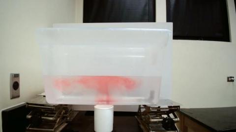 container of water with red food dye making a mushroom cloud shape with the container propped up by four jacks and a cup of water underneath